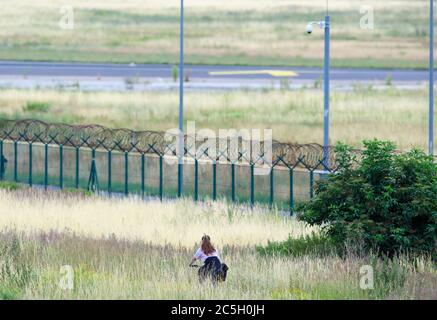 02 juillet 2020, Brandebourg, Schönefeld: Une jeune femme fait le tour de son vélo parallèle à la clôture de l'aéroport à travers l'herbe haute. Photo: Soeren Stache/dpa-Zentralbild/ZB Banque D'Images