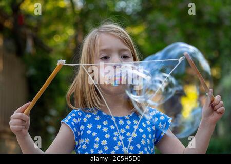 Jeune fille dans une robe bleue et blanche soufflant de grandes bulles avec une corde dans une cour arrière Banque D'Images