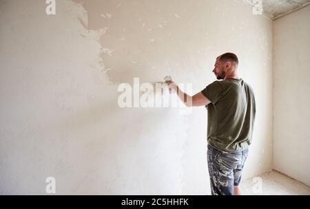 Vue arrière de l'ouvrier masculin fait la réparation dans l'appartement en mettant le mastic sur les murs. Jeune homme avec une barbe dans un T-shirt et des shorts tachés de peinture travaille jour. Maison de construction. Banque D'Images