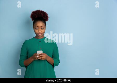 Téléphone intelligent à défilement. Portrait de la jeune femme afro-américaine isolé sur fond de studio bleu. Magnifique modèle féminin. Concept des émotions humaines, expression faciale, ventes, publicité. CopySpace. Banque D'Images