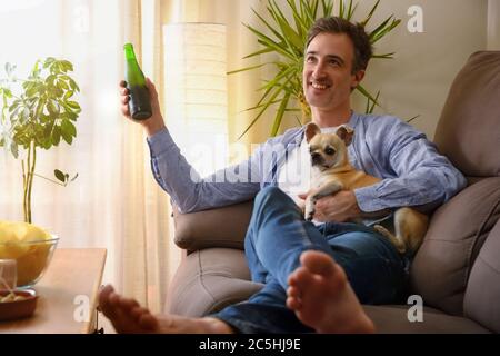 Homme souriant au repos ayant un snack assis sur un canapé à la maison avec son chien dans les bras Banque D'Images