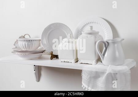 Composition d'une étagère suspendue en bois blanc sur le mur avec vaisselle maison de campagne en démonstration. Plaques à motif romantique, boîtes de rangement en céramique. Banque D'Images