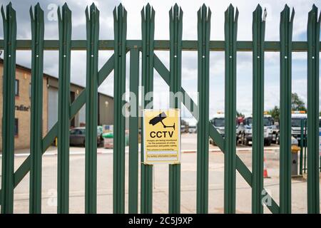 Mise au point peu profonde d'un panneau de sécurité de vidéosurveillance visible sur une clôture industrielle en métal et sécurisée. Au-delà de la clôture se trouve un grand entrepôt et des immeubles de bureaux. Banque D'Images