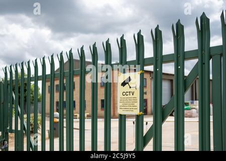 Mise au point peu profonde d'un panneau de sécurité de vidéosurveillance visible sur une clôture industrielle en métal et sécurisée. Au-delà de la clôture se trouve un grand entrepôt et des immeubles de bureaux. Banque D'Images