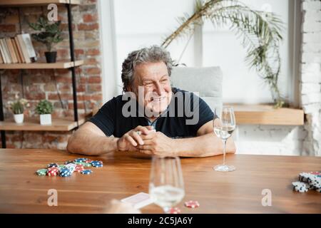 Passionnant. Joyeux homme mature jouant des cartes et buvant du vin avec des amis. Il a l'air ravi, enthousiaste. Homme caucasien jouant à la maison. Émotions sincères, bien-être, expression faciale concept. Bon vieux. Banque D'Images