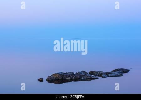 Paysage minimaliste tranquille avec des rochers sur la surface lisse du lac avec eau calme avec horizon avec ciel bleu clair au crépuscule, simple natura Banque D'Images