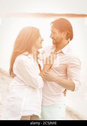 Couple souriant embrassant par la mer. Vue latérale de l'homme et de la femme en vêtements blancs se regardant l'un l'autre sur la plage de sable. Image teintée Banque D'Images