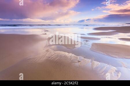 Après le lever du soleil orageux sur la côte, le dernier des nuages menaçants commence à adoucir la lumière et à créer des reflets sur le sable de la plage. Banque D'Images