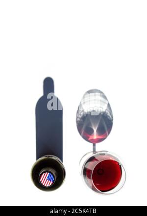 Une bouteille de vin et un verre de vin rouge jettent des ombres dures sur une table blanche. Le drapeau des États-Unis est représenté sur le bouchon de la bouteille. Banque D'Images