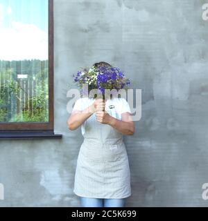 La fille dans un tablier cache son visage derrière un bouquet de fleurs de prairie. La fenêtre reflète le ciel et la clôture. F Banque D'Images