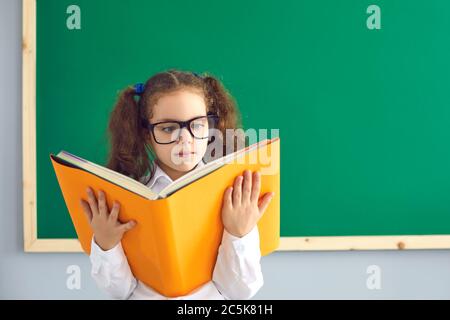 Retour à l'école. Smart petite fille dans des verres lisant un énorme livre près du tableau noir à la salle de classe. Adorable élève étudiant à l'école Banque D'Images