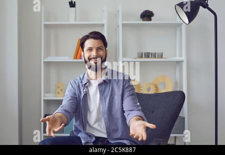 Jeune homme caucasien regardant la caméra chat vidéo avec les bras ouverts dans un geste amical de bienvenue dans le salon. Banque D'Images