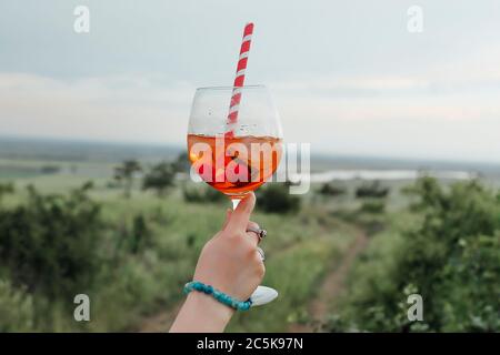 Femelle anonyme portant un gobelet de cocktail d'alcool avec des cerises et de la paille rayée sur fond flou de campagne Banque D'Images