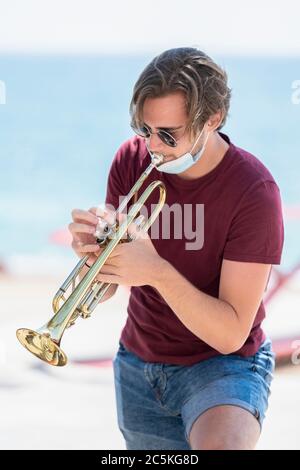 Beau jeune homme portant des lunettes avec son masque chirurgical tiré en jouant une trompette à l'extérieur sur un fond hors foyer. Musique et nouvelle vie Banque D'Images