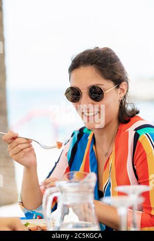 Souriante, belle jeune femme porte des lunettes tenant une fourchette avec de la nourriture sur un fond défocayeux. Manger à l'extérieur et concept de vacances. Banque D'Images
