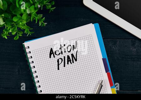 Vue du dessus du carnet ouvert écrit avec inscription AU PLAN D'ACTION. Fleur verte, tablette, stylo et carnet sur fond de bois. Banque D'Images