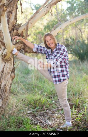 Portrait actif et femme sportive mature s'étirant dans la forêt, avec une branche d'arbre, heureux joyeux et détendu souriant, nature australienne Banque D'Images