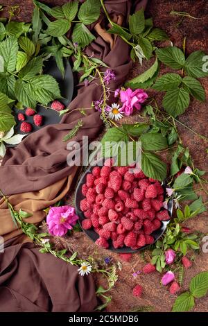 Framboises fraîches juteuses sur une assiette noire. L'été encore la vie avec, framboises, fleurs et herbes de prairie. Vue du dessus. Banque D'Images