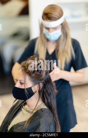 Coiffeur, protégé par un masque, peignant les cheveux de son client avec un fer à repasser dans un salon. Banque D'Images