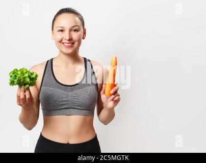 une jeune femme mince, avec une belle figure, tient des légumes dans ses mains - carottes et laitue sur fond blanc. Banque D'Images