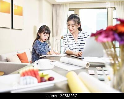 jeune femme de créateur asiatique travaillant à domicile à l'aide d'un ordinateur portable tout en prenant soin de sa fille (œuvres d'art en arrière-plan modifiées numériquement) Banque D'Images