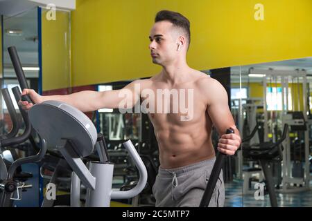 Jeune homme musclé s'exerçant sur machine elliptique, à l'intérieur d'une salle de gym. Banque D'Images