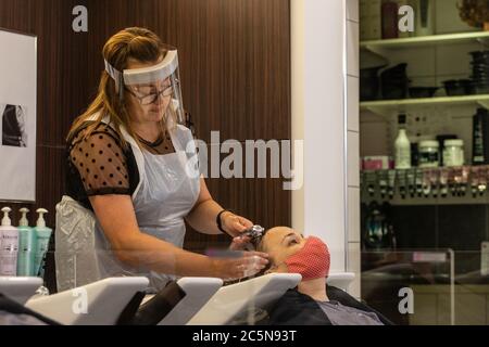 Coiffeur portant un EPI visage visière laver les cheveux du client portant un masque dans un salon de coiffure pendant les premiers jours de l'assouplissement du verrouillage en Angleterre 2020 Banque D'Images