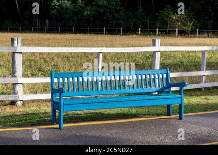 banc en bois bleu sur le côté d'une route asphaltée avec des marques jaunes en arrière-plan un cimetière avec un pré d'herbe sèche clôturé par une clôture. Banque D'Images