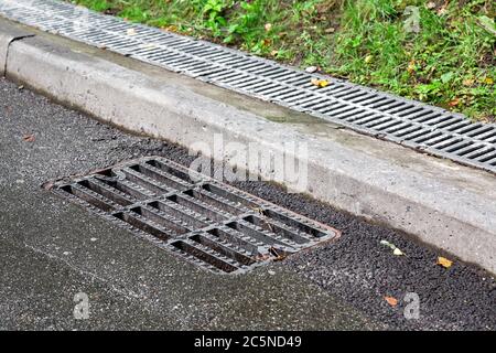 grille de trou d'homme d'égout sur une route asphaltée humide après la pluie près du trottoir en arrière-plan derrière le trottoir un canal d'évacuation recouvert d'une grille. Banque D'Images