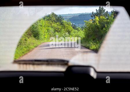 Route asphaltée parmi les arbres de la forêt. Vue de l'intérieur de la cabine par la lunette arrière d'une voiture sale. Banque D'Images