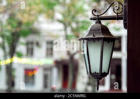 lanterne à charnières en fer avec inserts en verre accrochés sur un poteau contre le fond du bâtiment et arbres en effet de flou artistique. Banque D'Images