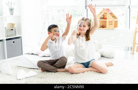 Des enfants heureux avec des lunettes sur des bâtons levant les mains assis sur le sol dans une pièce lumineuse Banque D'Images