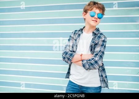 Portrait de mode de cheveux blancs blond jeune garçon de 12 ans t-shirt habillé et chemise à carreaux en lunettes de soleil bleues posant sur le bac bleu turquoise Banque D'Images