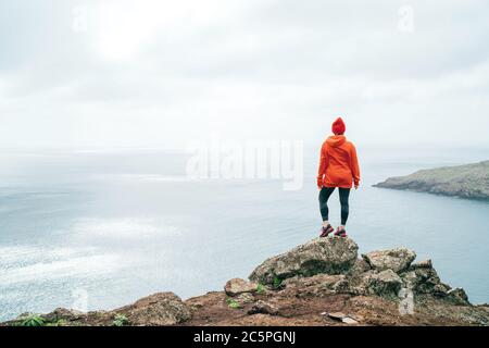 Trail Runner femelle vêtue orange sport sweat à capuche et rouge cap reposant sur la falaise et profitant de la vue sur l'océan Atlantique sur la péninsule de Ponta de Sao Lourenço Banque D'Images