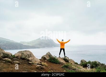 Trail runner homme habillé orange veste imperméable, collants de course et chaussures profitant de la vue sur la baie de l'Atlantique sur la péninsule de Ponta de Sao Lourenço - le e Banque D'Images