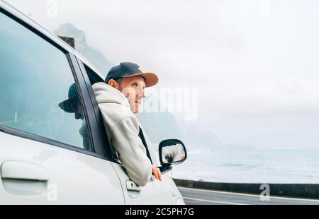 Homme habillé chaud sweat à capuche et casquette de baseball conduisant sa nouvelle voiture sur la route solitaire menant par la côte de mer. Image de concept de propriétaire d'auto heureux. Banque D'Images