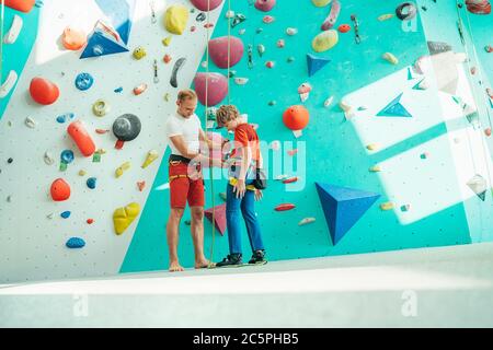 Père aidant son fils adolescent et vérifiant le harnais d'escalade près du mur d'escalade intérieur. Garçon se préparant pour une escalade. Concept de parent heureux imag Banque D'Images