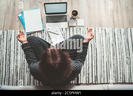 Femme d'affaires vêtue de pyjama méditant avec le café du matin avec des palmiers Gyan Mudra.sur le bureau d'étage avec ordinateur portable, les papiers vue de dessus. Distance Banque D'Images