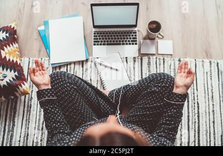 Femme d'affaires vêtue de pyjama, Ecoute d'écouteurs méditant avec le café du matin avec la paume Gyan Mudra au bureau avec ordinateur portable, les papiers vue sur le dessus Banque D'Images