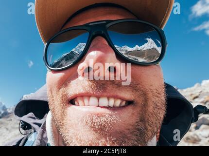 Photo en grand angle de l'objectif de la montagne en haute altitude, souriant, rasé, randonneur heureux en casquette de baseball avec des sommets enneigés et une chaîne montagneuse à côté de h. Banque D'Images