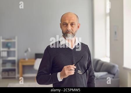 Portrait d'un homme âgé de race blanche à barbe grise regardant dans un appareil photo dans un arrière-plan de salon de maison moderne, tenant des lunettes. Concept de professionnel e Banque D'Images