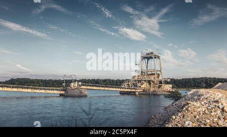 Tapis convoyeur industriel d'une installation d'extraction de gravier sur l'eau avec ciel sombre moody. Banque D'Images