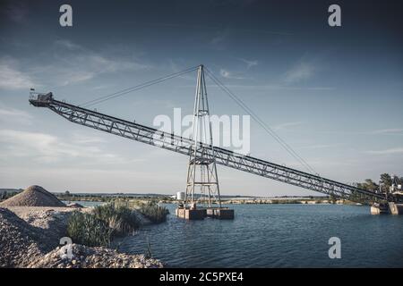 Tapis convoyeur industriel d'une installation d'extraction de gravier sur l'eau avec ciel sombre moody. Banque D'Images