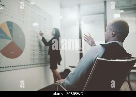 Vue de derrière un homme d'affaires dans une salle de réunion sur un fauteuil de bureau observant comment sa collègue femme d'affaires dans un arrière-plan défoqué Banque D'Images