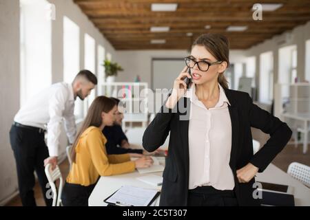 Jeune femme d'affaires sérieuse en lunettes et chemise parlant intelligemment sur téléphone portable au bureau avec des collègues sur fond flou Banque D'Images
