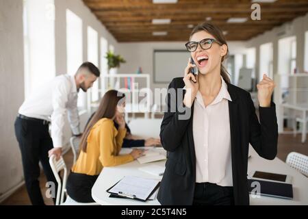 Une jeune femme d'affaires en lunettes et en chemise hurle tout en parlant au téléphone portable au bureau avec des collègues en arrière-plan Banque D'Images
