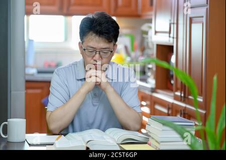 Homme asiatique d'âge moyen assis à un bureau à la maison, lisant un livre et étudiant. Banque D'Images
