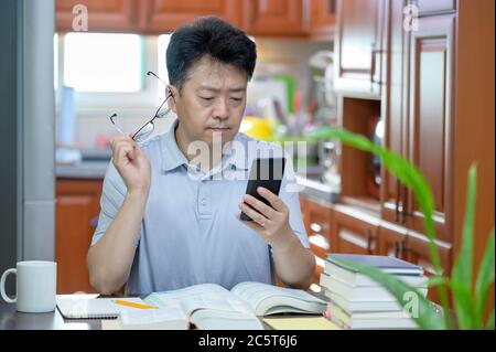 Homme asiatique d'âge moyen assis à un bureau à la maison, lisant un livre et étudiant. Banque D'Images