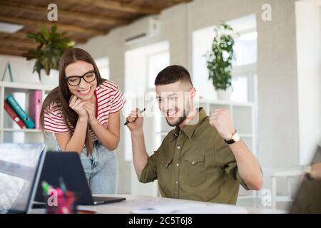 Jeune homme joyeux en chemise et femme en t-shirt rayé et lunettes de vue travaillant pleinement avec un ordinateur portable. Des gens d'affaires souriants et créatifs Banque D'Images