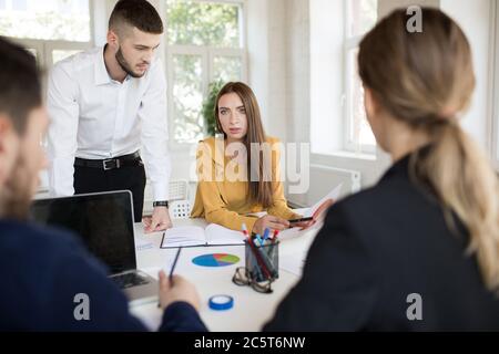 Belle femme d'affaires avec diagramme dans les mains regardant de côté tout en passant du temps au travail. Groupe de jeunes travaillant sur un nouveau projet Banque D'Images